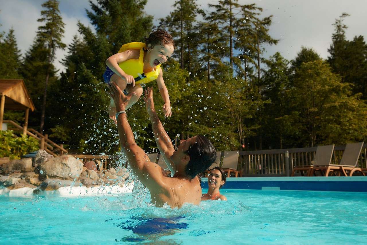 Child in a yellow life jacket laughs as an adult lifts them above water; another adult smiles nearby in an outdoor pool bordered by lounge chairs, railing, and tall trees.
