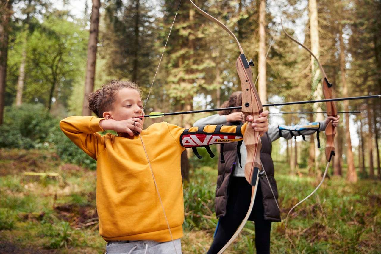 Two cyclists ride along a winding paved path through a sunlit forest, wearing helmets; the front rider smiles, trees and grass line the route, dappled light filtering through leaves.
