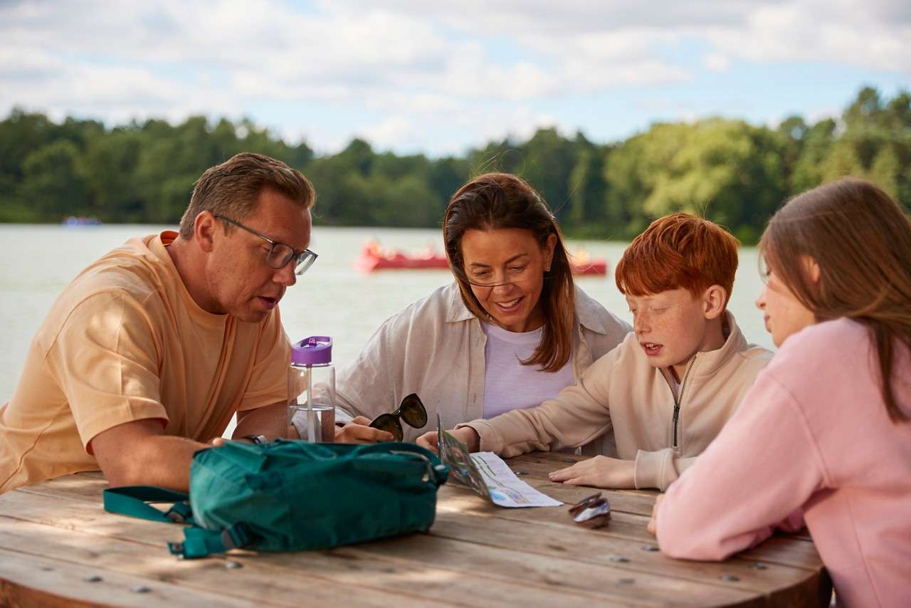 Child in a yellow life jacket laughs as an adult lifts them above water; another adult smiles nearby in an outdoor pool bordered by lounge chairs, railing, and tall trees.