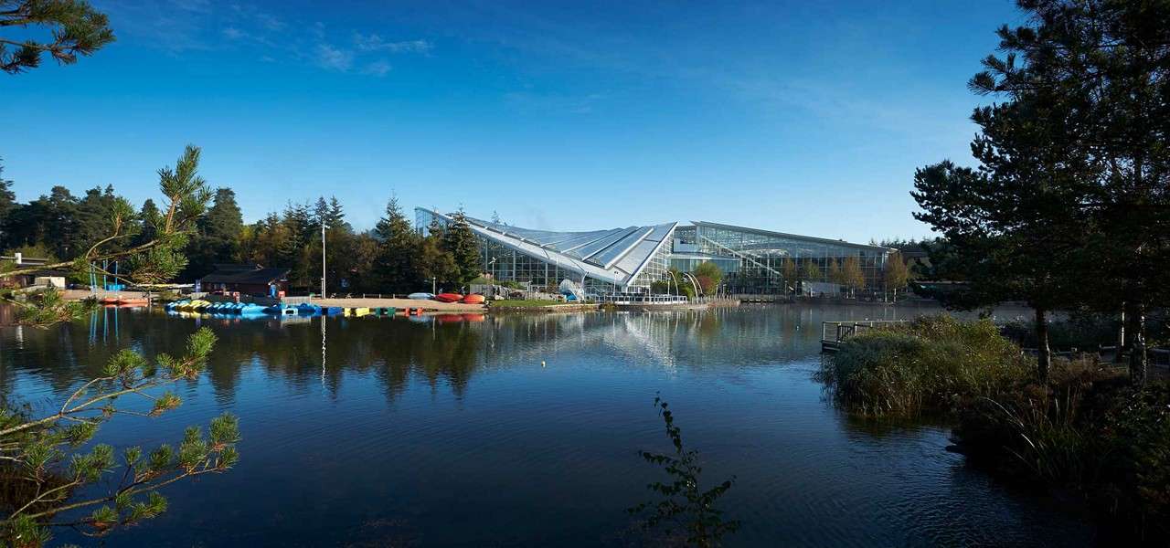 Glass-roofed leisure complex reflects on a calm lake; colorful kayaks and pedal boats rest at the dock. Pine trees frame the scene under a blue sky in a forested park.