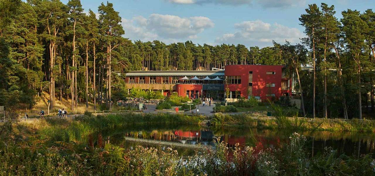 A modern red building reflects in a calm pond, as visitors walk and cycle nearby, nestled among tall pine trees under a partly cloudy sky within a landscaped park.