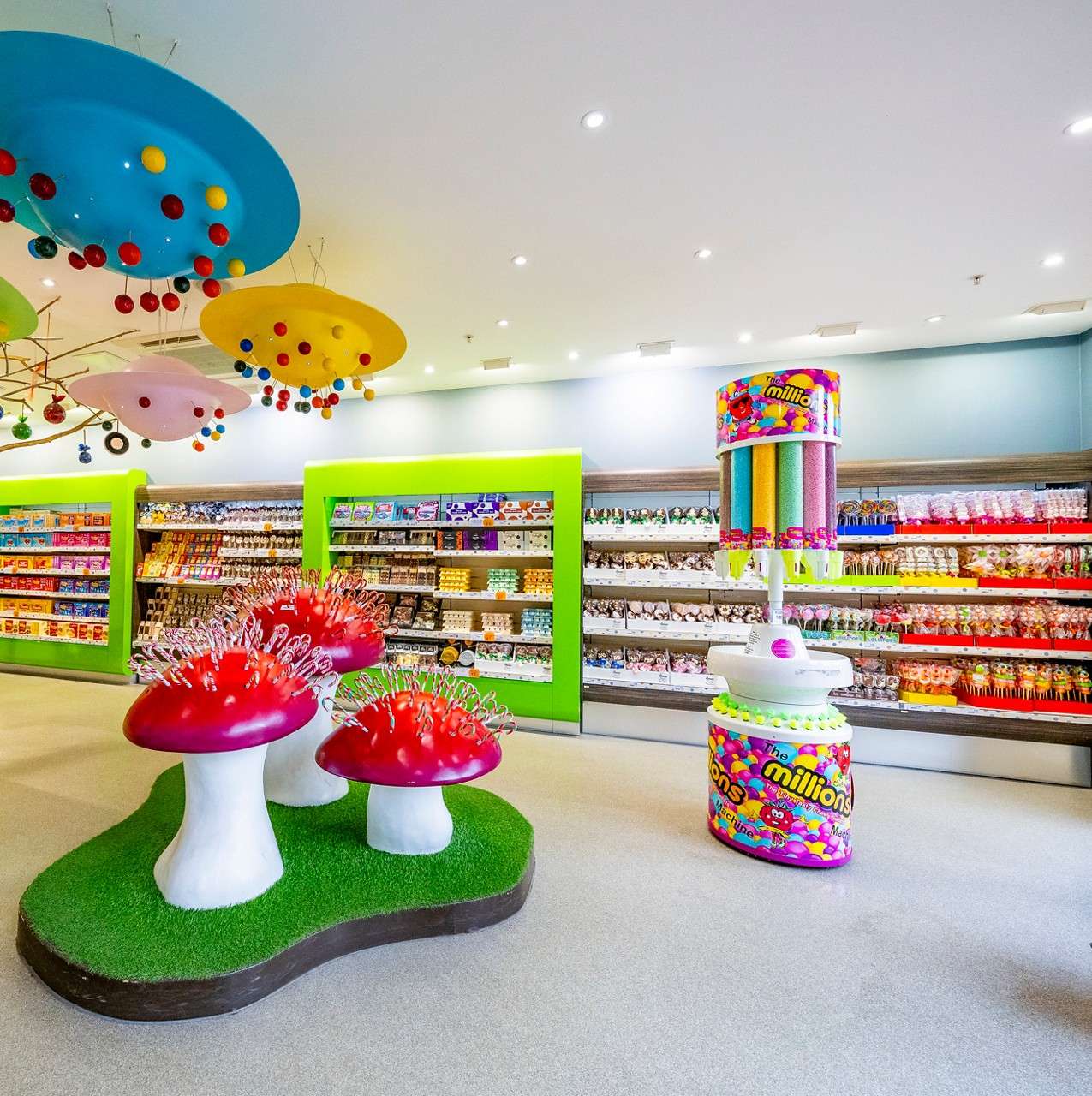 Candy aisles display assorted sweets, while a colorful dispenser and giant red-capped mushroom sculptures enliven a bright, playful store with ceiling mobiles. Text: “the millions”.