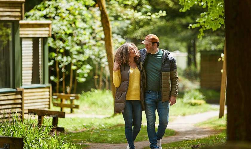 Two adults walk arm-in-arm, smiling, along a curving path; surrounding wooden cabins, benches, and lush green trees suggest a quiet forest resort on a sunny day.