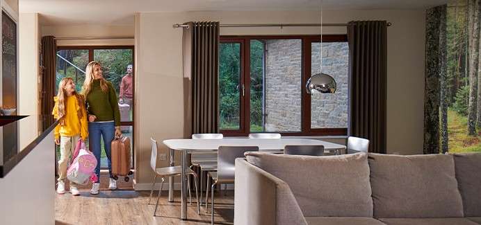 Family with luggage enters a modern vacation cabin; a woman and child carry bags while a man follows outside. Around them, a dining table, chairs, sofa, big windows, forest view.