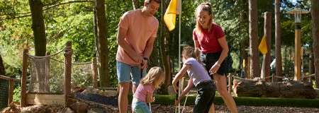 Two adults and two children play miniature golf, bending with putters near a hole. Sunlit forest setting surrounds them, with yellow flags, wooden obstacles, rope fence, and gravel paths.