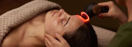 Face receives treatment; a handheld device emits red light onto the forehead while the person lies under a textured blanket on a spa bed, head supported by a practitioner.