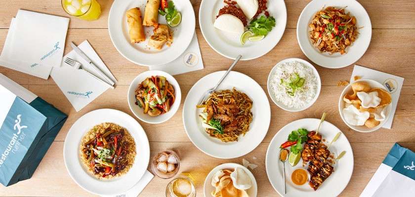 Assorted dishes sit on a wooden table; noodles, rice, skewers, steamed buns, salads, sauces, drinks, and prawn crackers surround takeout bags and napkins.

Text: “restaurant” (repeated on napkins, bag, and sticker; smaller words illegible).