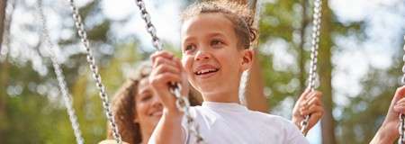 Child grips swing chains and smiles while swinging; adult behind assists. Sunlit park with blurred trees forms background.