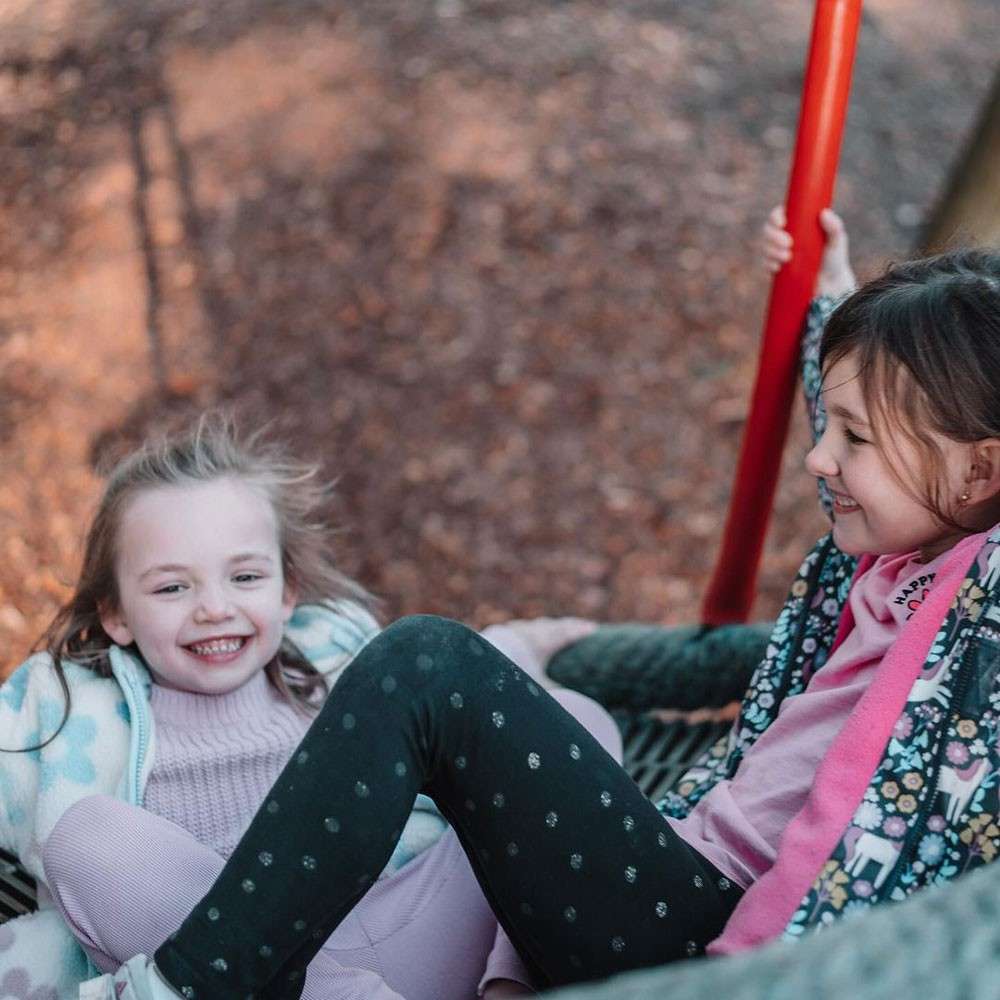 Two children laugh while sitting together on a net swing; one grips a red pole. They wear patterned jackets and scarves. Background shows blurred brown leaves. Visible text: HAPPY.
