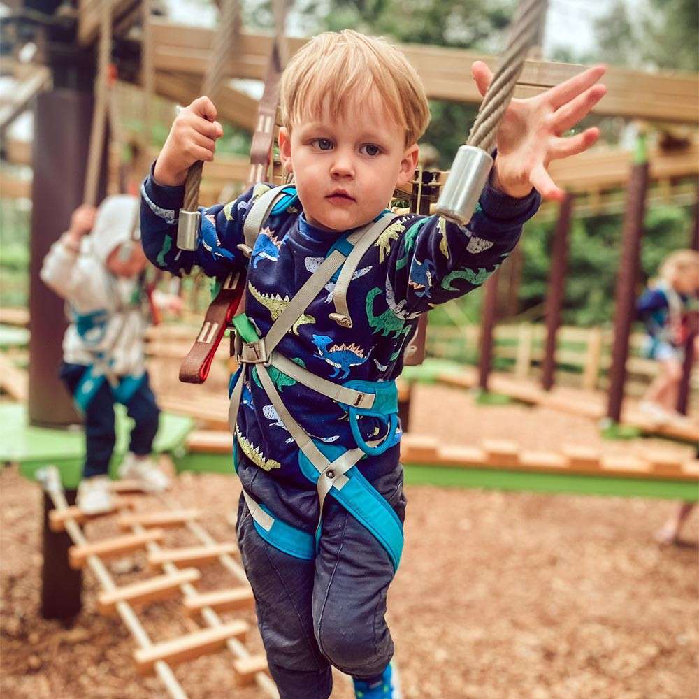 Child in safety harness reaches for a rope, stepping across a suspended obstacle. Context: outdoor adventure playground with woodchip ground, rope ladders, wooden platforms, and other children in the background.