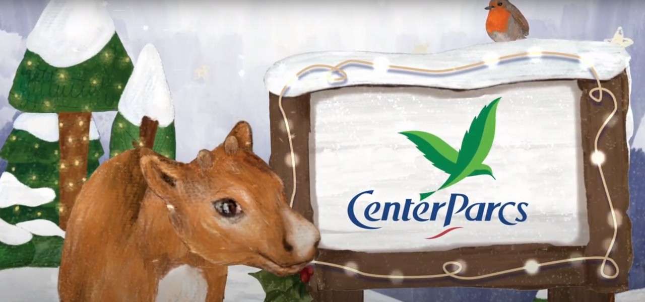 A deer gazes at a snowy wooden sign displaying the green-bird logo and text Center Parcs; a robin perches atop, with string lights and snow-covered pine trees in the background.