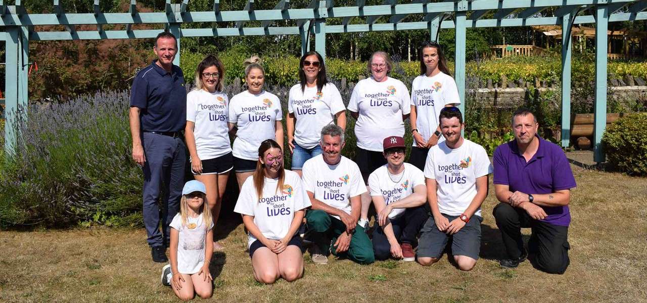 Group of volunteers pose and kneel in a sunny garden under a pergola. Visible text: "together for short LIVES" (on multiple shirts), "NY" (on cap).