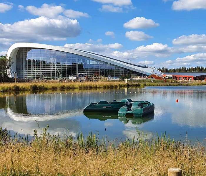 Pedal boats float on a calm pond, reflecting a large curved glass-fronted building. Grasses line the shore; a red buoy drifts under a bright, partly cloudy sky with distant trees.