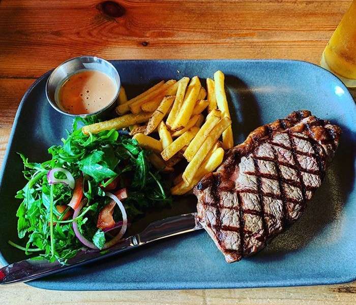 Grilled steak sits beside golden fries and a mixed green salad with tomato and red onion, plus a small cup of sauce, on a gray plate atop a wooden table.