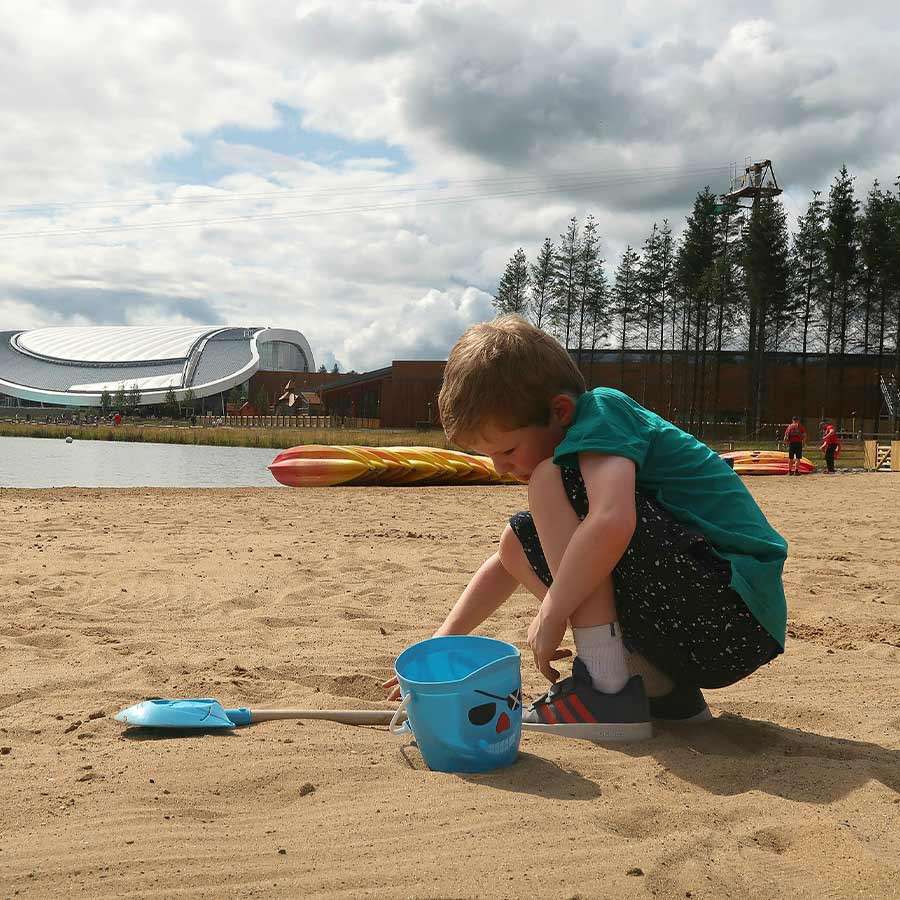 Child crouches and digs in sand with a small blue bucket and spade, beside a lake; background shows modern curved-roof building, pine trees, scattered people, and cloudy sky.
