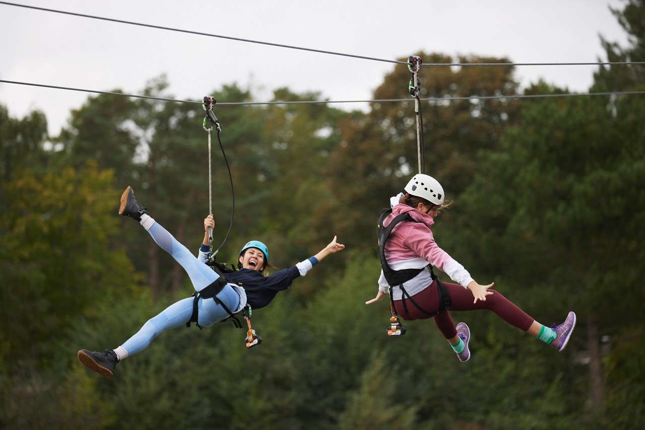 Two helmeted riders glide side-by-side on zip lines, legs splayed and hands gripping tethers, suspended by harnesses above a wooded landscape on an overcast day.