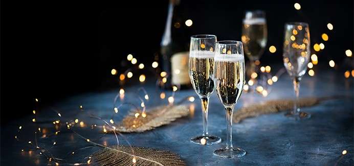 Four glasses of champagne on a light blue table decorated with twinkling lights and bronze leaf decorations. Bottle of champagne sits on the table in background. Two glasses are in focus in the dark room.