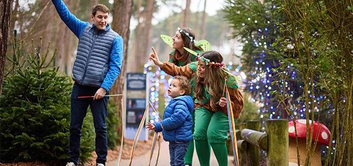 Adult holding a bundled child gazes upward, standing inside a tunnel of blue-green string lights, both wearing knit beanies and winter coats in a nighttime display.