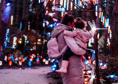 Child and adult sat at a red table painting a wooden reindeer, smiling whilst looking at each other. Child is wearing elf hat and elf costume. Christmas tree is adorned with multicoloured lights twinkling in the background.