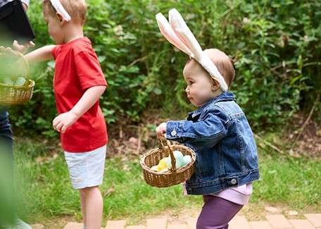 Child and adult sat at a red table painting a wooden reindeer, smiling whilst looking at each other. Child is wearing elf hat and elf costume. Christmas tree is adorned with multicoloured lights twinkling in the background.