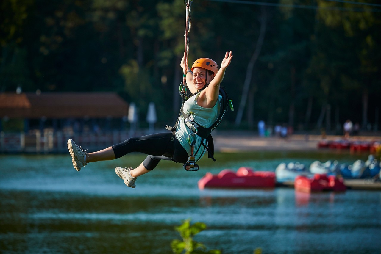 Person in helmet and harness zip-lines with arms raised, legs extended, smiling. Over a lake, with paddle boats, dock, and forested shoreline in the background.