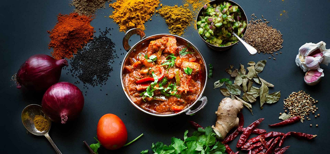 A bowl of red curry sits garnished with cilantro and chilies, surrounded by onions, garlic, ginger, curry leaves, chilies, cumin and coriander seeds, and spice piles on a dark surface.