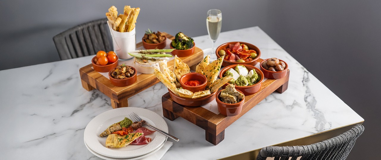 Assorted tapas bowls sit on wooden risers, presenting breads, cheeses, olives, mushrooms, vegetables, and dips; a plate with samples and fork rests on a marble table beside a champagne flute.