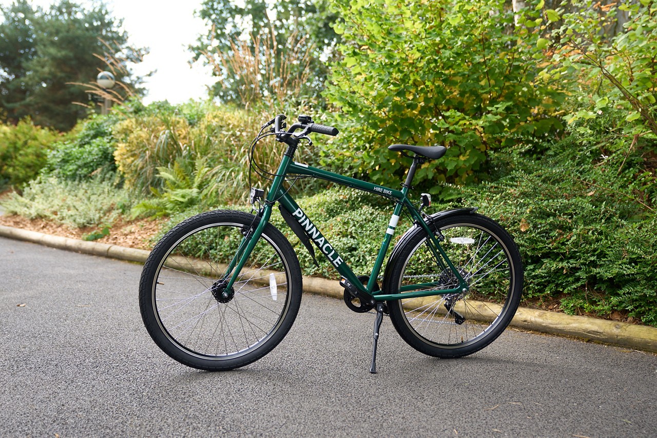 Bicycle stands on its kickstand, resting on an asphalt path beside lush garden shrubs and trees. Visible text on frame: PINNACLE; HIRE BIKE.