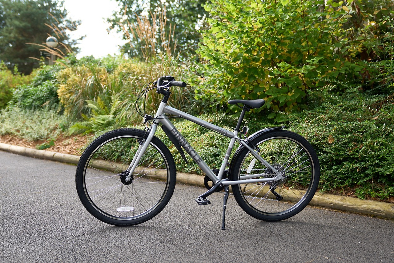 Silver hybrid bicycle stands on its kickstand, parked on a paved path; leafy shrubs and trees surround it. Visible text: PINNACLE.