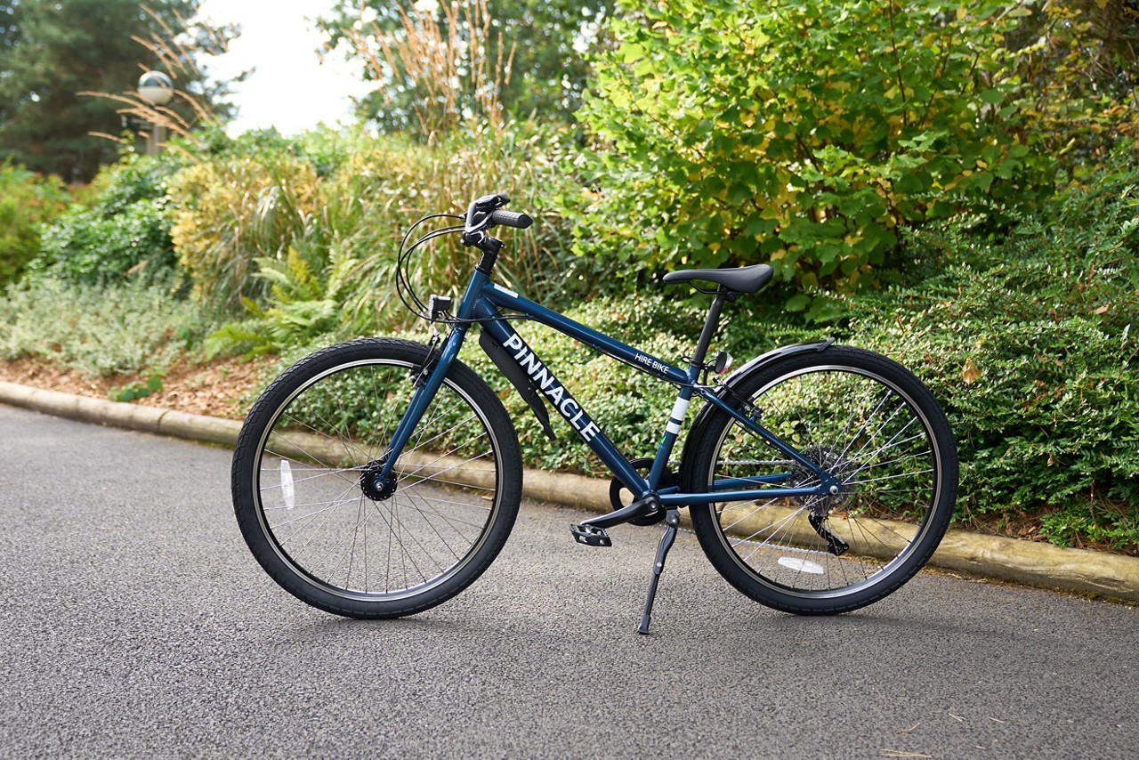 Bicycle stands on its kickstand, facing left, on a paved path beside dense greenery and a curb. Frame text: PINNACLE, HIRE BIKE.