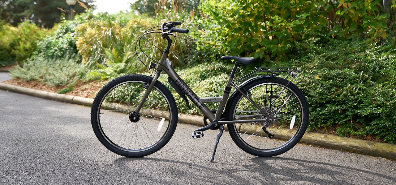 Commuter bicycle stands on its kickstand, facing left, with rear rack and fenders; context: sunlit paved path bordered by dense green shrubs and trees. Text on frame: HIRE BIKE.