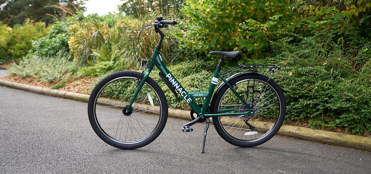 Green step-through bicycle stands on its kickstand, with rear rack and fenders, on a paved path beside dense shrubs; text on frame: PINNACLE and HIRE BIKE.