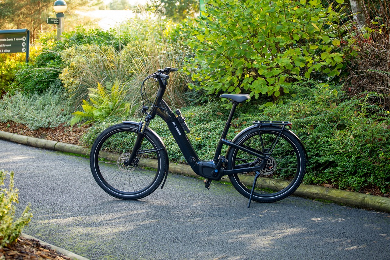 Electric bicycle stands on its kickstand, resting on a paved path, surrounded by shrubs and trees; step-through frame, fenders, disc brakes, rear rack. Text: SPECIALIZED.