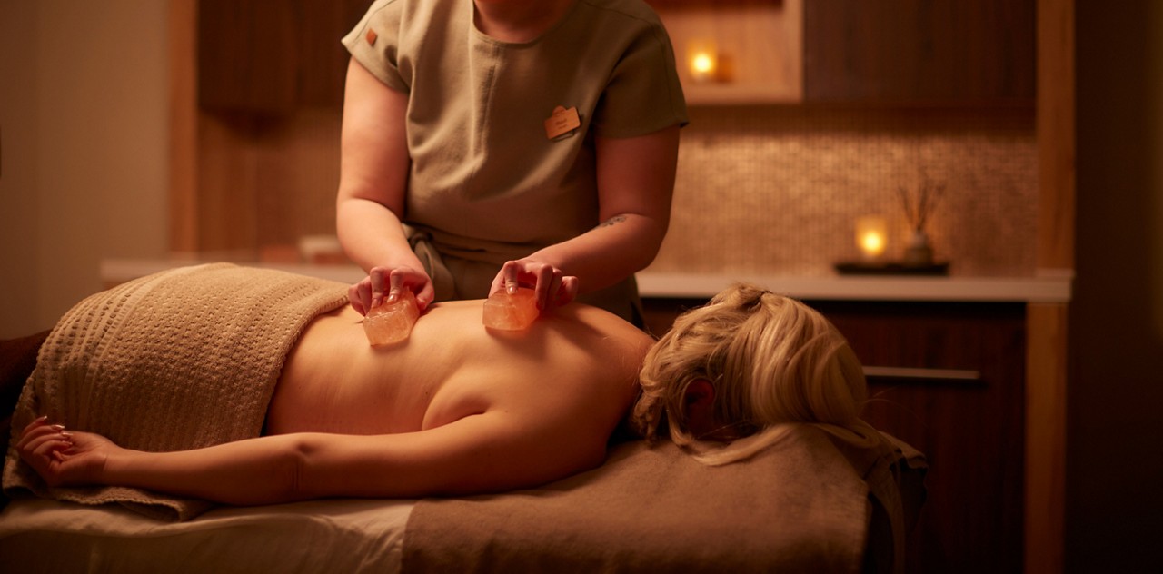 Massage therapist glides two warm stones across a client’s bare back. Dimly lit spa room surrounds them with soft candles, wooden cabinets, towels, and a calming, warm-toned ambiance.