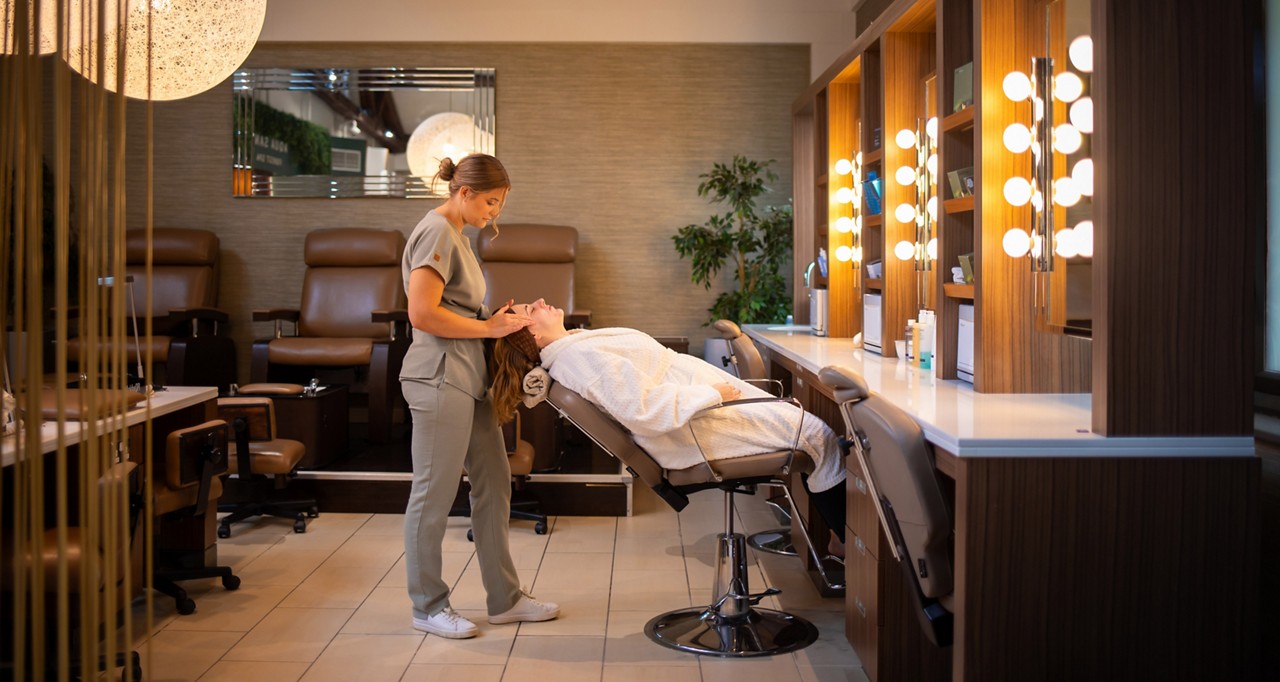 Esthetician massages a client’s face while the client reclines in a salon chair; illuminated vanity mirrors, wood cabinetry, and cushioned seating define a calm, professional spa environment in the background.