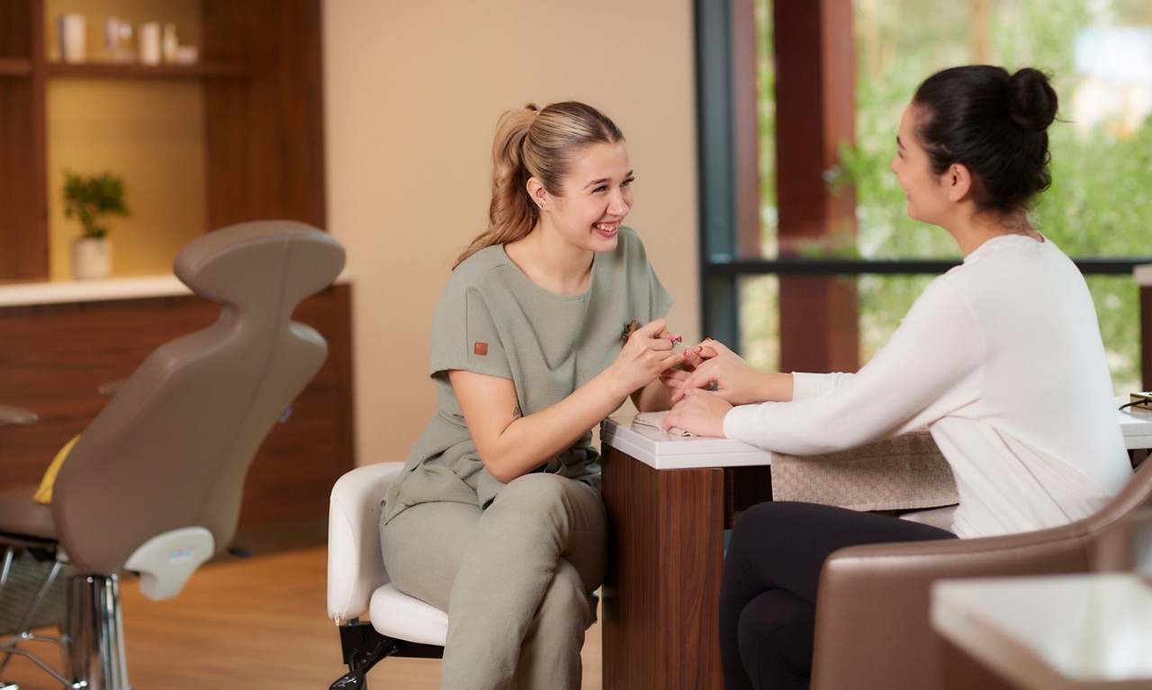 Manicurist trims client’s nails at a salon table; both women smile and chat. Context: modern, well-lit spa with cushioned chairs, wooden shelves, potted plant, and large window showing greenery.