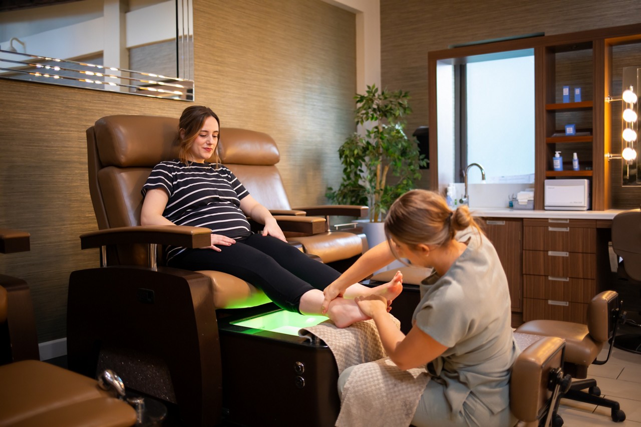 Client reclines on a pedicure chair, receiving a foot massage from a technician, in a modern spa with brown leather seats, glowing green footbath, shelves, sink, and vanity lights.