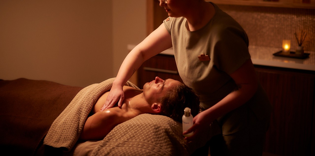 Client lies on a massage table, receiving a chest and shoulder massage from a therapist applying oil. Dim spa room with blankets, cabinets, and a lit candle in the background.