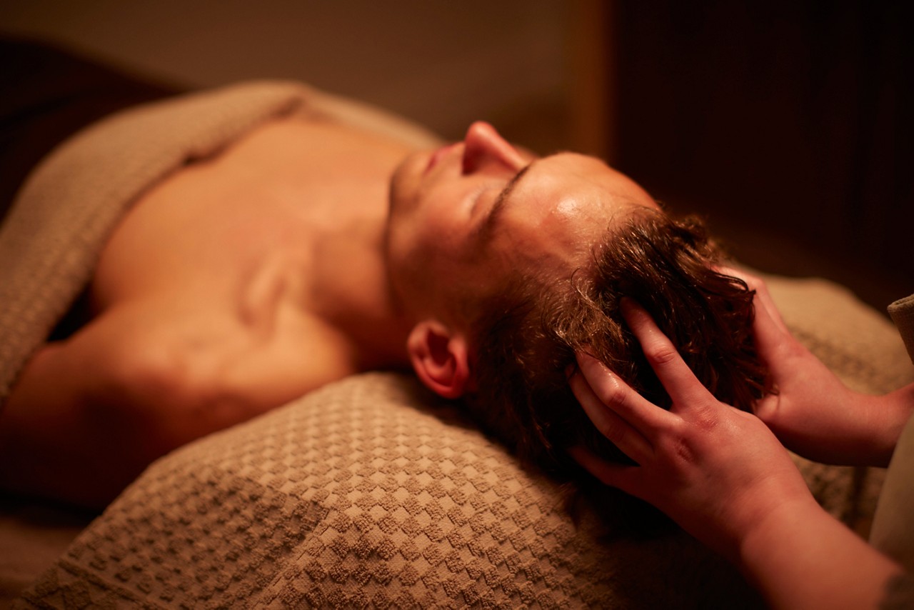 Person receives scalp massage while lying on a massage table; therapist's hands knead hair; warm dim lighting and textured towels create a calm spa setting.
