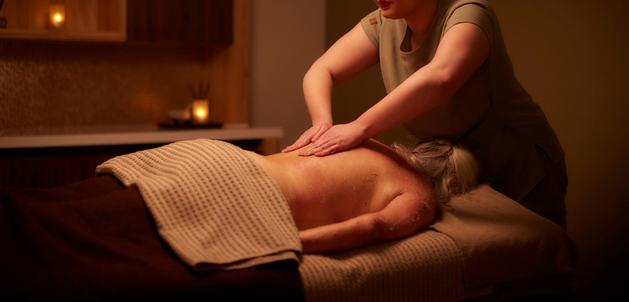 Client lying face down is massaged; therapist presses both hands on bare back. Dim spa room with candles, towels, and a textured blanket on the table.