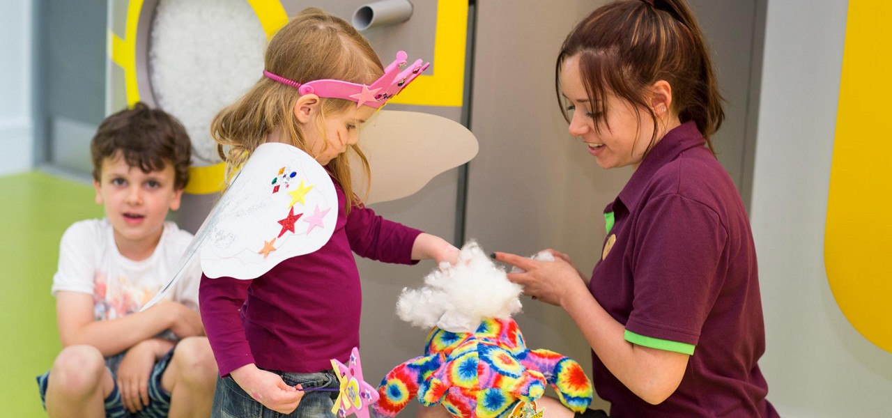 Child wearing fairy wings fills a colorful teddy bear with stuffing, assisted by a worker; another child watches in a bright workshop with a stuffing machine and playful decor.