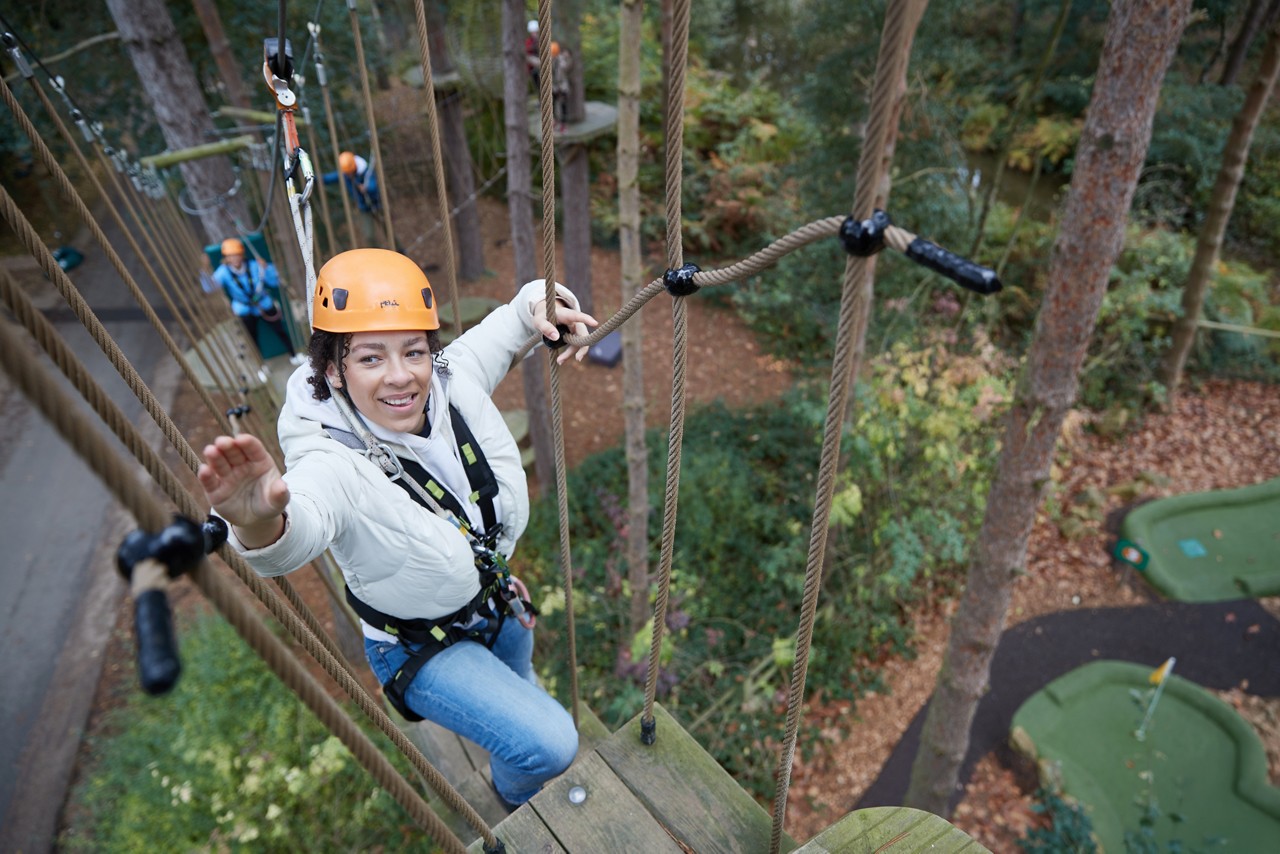 Person wearing orange helmet and harness crosses a rope bridge, reaching for a handhold. Set in a treetop adventure course amid tall trees, with participants and platforms visible below.